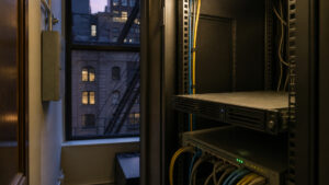Dim NYC office closet with a leftover on-premises mail server and a laptop showing a disconnected hybrid path, no readable text.