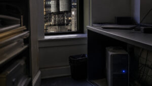Dim NYC office nook with a leftover tower PC under a desk and water towers outside the window, suggesting an unpatched on-premises SQL Server.