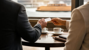 Two professionals exchanging a business card at a NYC cafe table with a ferry visible through the window, suggesting a warm referral introduction.
